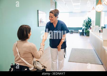 Infermiera sorridente che saluta paziente seduta su sedia a rotelle con stretta di mano nella hall dell'ospedale Foto Stock