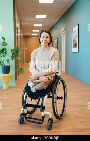 Ritratto di donna matura che ha disabilità fisica seduta su sedia a rotelle nella hall dell'ospedale Foto Stock
