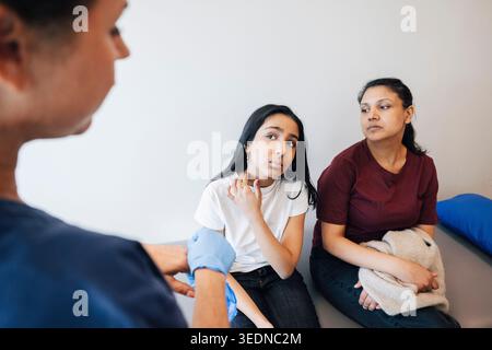 Vista ad alto angolo della madre seduta accanto alla figlia che mostra problemi al collo a una donna esperta di assistenza sanitaria in sala esame Foto Stock