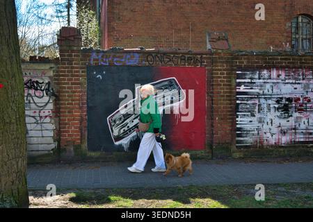 Donna anziana che cammina con il cane piccolo, supera il murale della cassetta dei graffiti sul muro di mattoni urbani Foto Stock