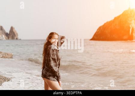 Donna da spiaggia flanella donna serena in una camicia a quadri oversize in piedi sulla costa con onde oceaniche e scogliere rocciose durante un tranquillo tramonto Foto Stock