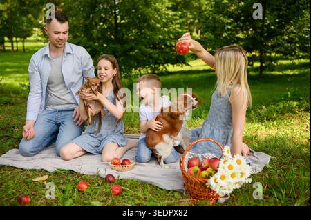 Famiglia felice con bambini e cani che si divertono al picnic nel parco estivo. Genitori e bambini che giocano con animali domestici su erba verde. Concetto di infanzia felice, s Foto Stock