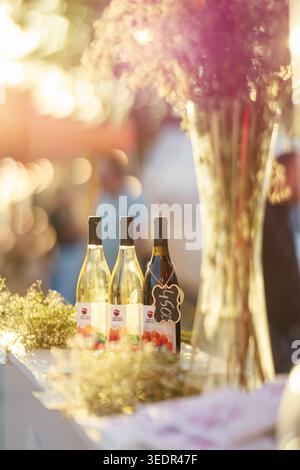Al Festival del vino di Yerevan, le bottiglie di vino marchiate vengono esposte su un bancone del festival accanto a un alto allestimento floreale nella calda luce serale di Yerevan. Foto Stock