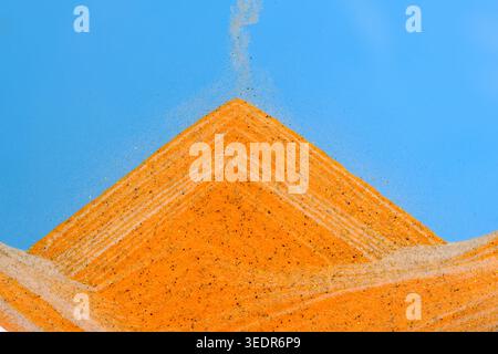 Sabbia arancione a triangolo perfetta che viene versata contro un cielo blu brillante, mostrando movimento e granulosità Foto Stock