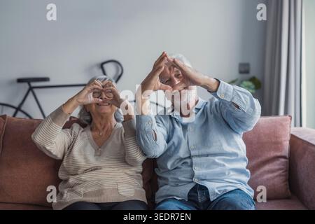 Una coppia anziana amorevole sta insieme in casa facendo un gesto cardiaco, esprimendo compassione, impegno e felicità condivisa in un ritratto intimo ed emotivo Foto Stock