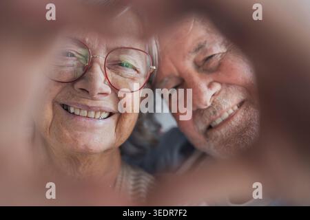 Una coppia in pensione felice posa in casa facendo un gesto cardiaco con le dita, sorridendo alla fotocamera ed esprimendo amore, sincerità e un forte legame emotivo in un ritratto caldo Foto Stock