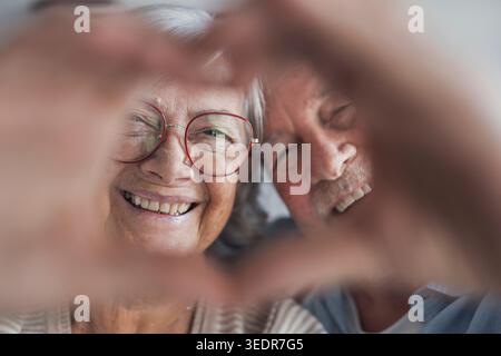 Primo piano ritratto felice sincero anziano di mezza età pensionato coppia di famiglia facendo gesto cardiaco con le dita, mostrando l'amore o dimostrando sentimenti sinceri insieme in interni, guardando la fotocamera. Foto Stock