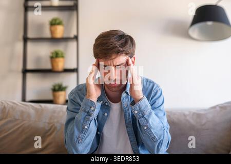 Un millennial stanco si siede su un divano a casa, toccando la testa con dolore mentre lotta con un forte mal di testa o emicrania, sperimentando affaticamento, mancanza di sonno, vertigini e problemi di vista dopo lunghi giorni di stress mentale Foto Stock
