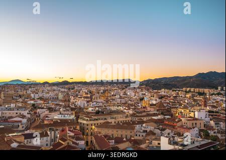 Ampia vista panoramica sulla città di Malaga, in Spagna, che mostra i fitti edifici urbani e i tetti con le colline circostanti sotto un colorato cielo al tramonto. Il Foto Stock