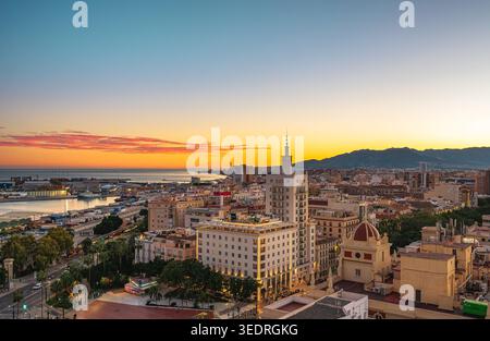 Ampia vista sopraelevata di Malaga, Spagna, che mostra lo skyline della città, il porto e il lungomare al tramonto, con montagne che si stagliano sotto un cielo limpido e colorato. St Foto Stock