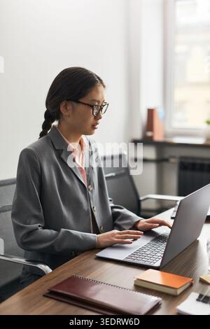 Immagine verticale di una giovane donna d'affari asiatica che lavora su un notebook alla scrivania dell'ufficio, gestisce attività online e comunicazioni mirate. Ideale per la produttività aziendale o il marketing da remoto Foto Stock