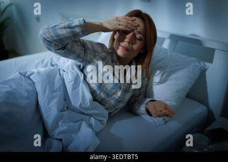 Una donna si siede a letto con la mano sulla fronte e tiene in mano la droga. Sembra a disagio mentre soffre di mal di testa di notte. Un bicchiere di Foto Stock