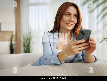 Una donna siede su un divano in una stanza luminosa e usa il suo telefono con un sorriso. Le piace il tempo libero mentre si rilassa. Foto Stock