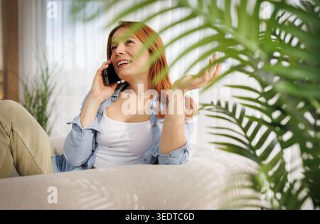 Una donna siede su un divano in un soggiorno. Sorride mentre parla al telefono. Le piante verdi sono sullo sfondo. Foto Stock