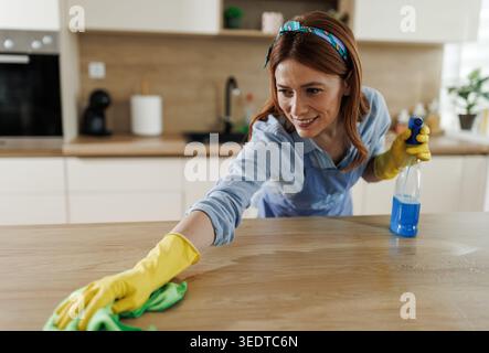 Una donna con guanti gialli utilizza un flacone spray e un panno per pulire la superficie della cucina. Si concentra sul suo compito in una cucina luminosa Foto Stock