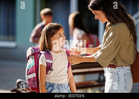 La gioiosa riunione ha luogo mentre la madre aspetta sua figlia dopo la scuola. Altri bambini giocano e socializzano in sottofondo nelle giornate di sole Foto Stock