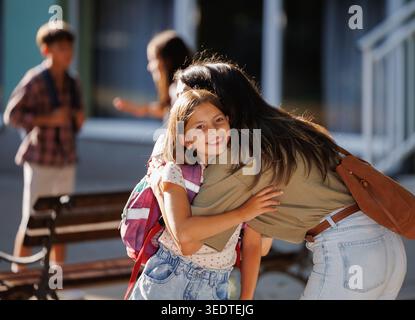 La gioiosa riunione ha luogo quando la madre abbraccia sua figlia dopo la scuola. Altri bambini giocano e socializzano in sottofondo nelle giornate di sole Foto Stock