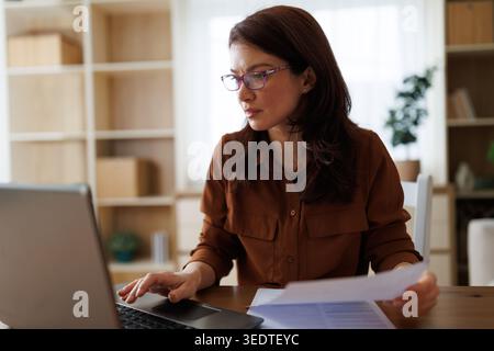 Una donna è concentrata sul suo lavoro al portatile mentre esamina i documenti. Si siede in una stanza luminosa con una semplice scrivania. Foto Stock