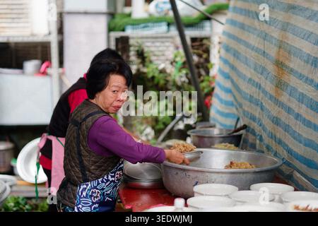 Taipei, Taiwan - 29 marzo 2025: Una cuoca anziana prende del riso appiccicoso da una grande ciotola mentre guarda la macchina fotografica durante il banchetto all'aperto a Neihu. Foto Stock