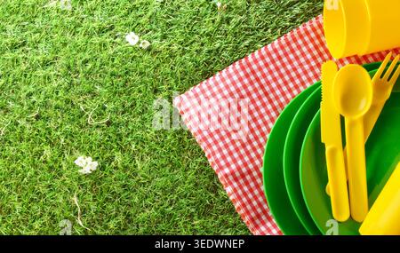 Vista dall'alto di colorati piatti da picnic e posate su una coperta a scacchi rossi sull'erba verde in un luminoso ambiente estivo all'aperto. Foto Stock