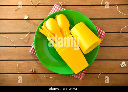 Posate e tazze in plastica di colore giallo brillante disposte su un piatto verde su un tavolo da picnic in legno con tovagliolo rosso a scacchi e fiori decorativi. Foto Stock