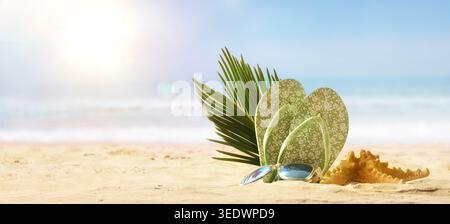 Infradito decorativo, occhiali da sole, foglie di palma e stelle marine che riposano su sabbia soffice con onde oceaniche e luce solare estiva brillante in una scena di spiaggia tropicale. Foto Stock
