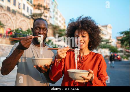 Giovani coppie che abbracciano la tendenza della cucina asiatica, mangiando felicemente spaghetti con bacchette all'aperto Foto Stock