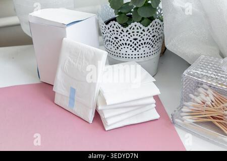Le confezioni di tessuto bianche sono posizionate ordinatamente su un tavolo accanto a una pianta in vaso e su un contenitore di tamponi di cotone. La scena mostra gli oggetti di tutti i giorni disposti in una casa spenta Foto Stock