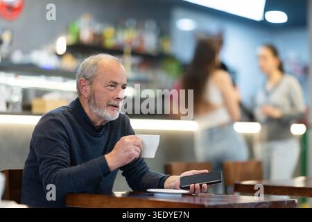 un uomo anziano beve caffè e tiene in mano un telefono cellulare Foto Stock