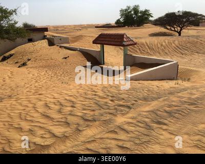 Vista degli edifici abbandonati e di un piccolo cancello con un tetto rosso parzialmente sepolto dalle dune di sabbia del deserto mutevoli a Dubai, Dubai, Emirati Arabi Uniti. Foto Stock