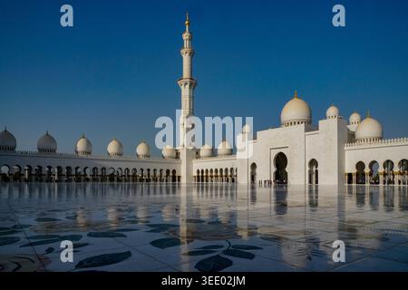 Vista della grande Moschea dello Sceicco Zayed con cupole in marmo bianco, un alto minareto e un cortile floreale riflettente sotto un cielo azzurro limpido ad Abu DH Foto Stock