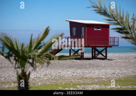 Capanna rossa di sicurezza in legno su palafitte situata su una spiaggia sassosa con foglie di palma in primo piano Foto Stock