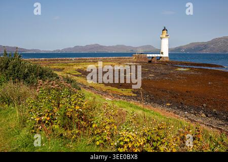 Veduta del Faro di Tobermory che sorge sulla costa rocciosa circondata da vegetazione verde e dalle acque blu del Sound of Mull con una collina lontana Foto Stock