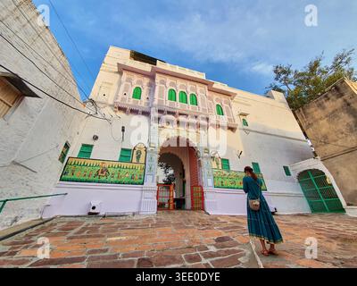 Una donna in un lungo vestito blu verde si trova in un cortile lastricato di pietra di fronte a un edificio tradizionale indiano haveli. La struttura imbiancata a calce presenta Foto Stock