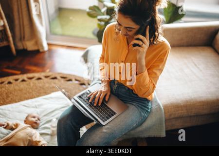 Una madre si siede sul divano di casa, utilizza un computer portatile e parla al telefono mentre un bambino riposa nelle vicinanze. Foto Stock