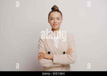 Immagine di una giovane e bella manager donna in abito beige e camicia bianca con le braccia incrociate, leggermente sorridente e guardando in avanti, pur avendo successo e sicurezza contro le pareti luminose Foto Stock