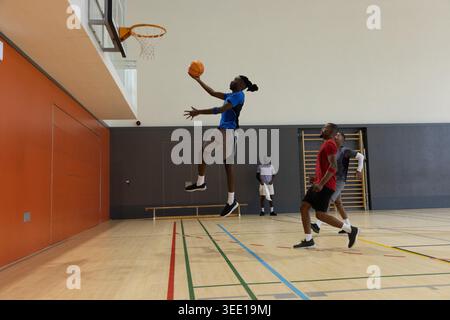 Diversi giocatori maschili di abbigliamento sportivo che giocano a basket in palestra, saltano a strapiombo verso il cerchio. Palla, campo, backboard, compagni di squadra, salto, perforazione dinamica Foto Stock