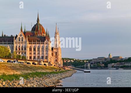 L'edificio del Parlamento si trova vicino al fiume a Budapest. La gente cammina lungo la riva mentre il sole tramonta nel cielo serale. Foto Stock