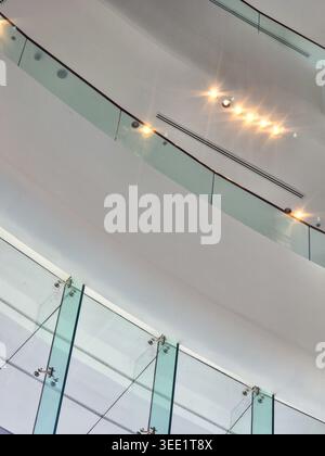 Moderno balcone in vetro con ringhiera, spazioso interno bianco dell'edificio che riflette la luminosa plafoniera a creare un'atmosfera aperta e un elegante design architettonico Foto Stock