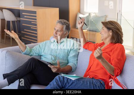 Coppia con donna anziana seduta sul divano di casa, uomo che tiene un tablet, donna che scrive su un notebook Foto Stock