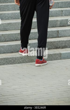 Sneakers rosse e pantaloni sportivi neri che camminano sul marciapiede urbano durante l'attività fisica all'aperto e il concetto di stile di vita sano Foto Stock