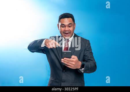 Ritratto di un dirigente indonesiano felice con un abito, che punta al suo smartphone con un'espressione sorpreso ed entusiasta in uno studio blu. Foto Stock