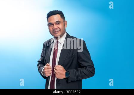 Ritratto di un dirigente indonesiano in tuta e cravatta, con la sua giacca in un'espressione sicura su uno sfondo blu da studio. Foto Stock