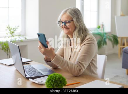 Donna anziana che lavora alla scrivania con un computer portatile che tiene in mano il telefono cellulare. Foto Stock