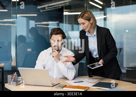 Due diversi colleghi aziendali che lavorano insieme su un progetto congiunto, discutono idee e analizzano i dati su un notebook e un tablet in un ambiente di ufficio contemporaneo Foto Stock