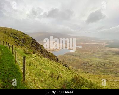 Vista panoramica a nord dal Conor Pass sui laghi glaciali e sulla valle bogland, sulla collina verde con recinzione a filo metallico, sulla penisola di Dingle, Kerry, Irlanda Foto Stock