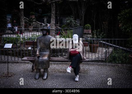 Un colpo sincero che cattura una donna con un berretto e un branco rosso di fanny seduto casualmente su una panchina del parco accanto a una statua di bronzo simile a un essere umano. L'impostazione è un'uscita Foto Stock