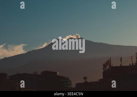 Vista distante del vulcano attivo dell'Etna, che mostra un pennacchio di fumo che sale dal suo cratere contro un cielo limpido durante il tramonto. Il featu in primo piano Foto Stock