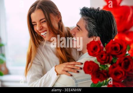 Giovane donna felice che ride mentre abbraccia il fidanzato dopo aver ricevuto rose rosse a casa, coppia romantica che celebra l'amore in un luminoso soggiorno moderno Foto Stock