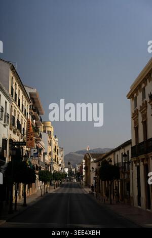 Strada vuota illuminata dal sole nella città andalusa con edifici bianchi e montagne lontane sotto il cielo blu. Foto Stock